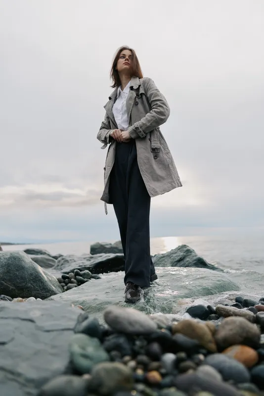 Young woman in a grey coat stands on rocky shore near ocean under cloudy sky. Casual fashion and nature concept with moody atmosphere and natural landscape background.