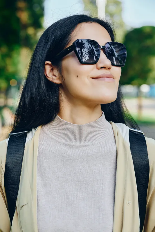 Sunglasses, woman outdoors backpack casual fashion park sunny confident streetwear style under natural light, relaxed pose conveying modern vibe and street style in a sunny outdoor setting