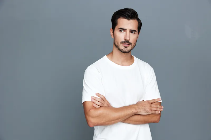 Thoughtful man with crossed arms wearing a white t-shirt looking to the side against a plain gray background in a studio portrait. Emotions concept.