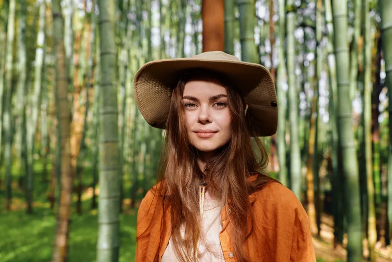 woman portrait wearing a wide straw hat in a bamboo forest, nature scene with warm sunlight and green trunks. Young adult model with long hair, calm gaze, casual boho jacket outdoors, relaxed