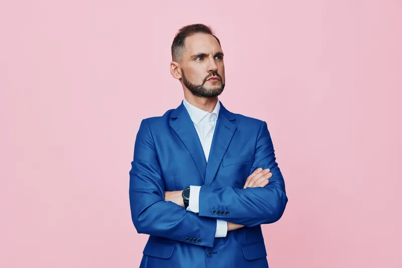 A professional male business figure stands with arms crossed against a solid pink background, conveying confidence and calm focus. The isolated color backdrop highlights his poised posture and