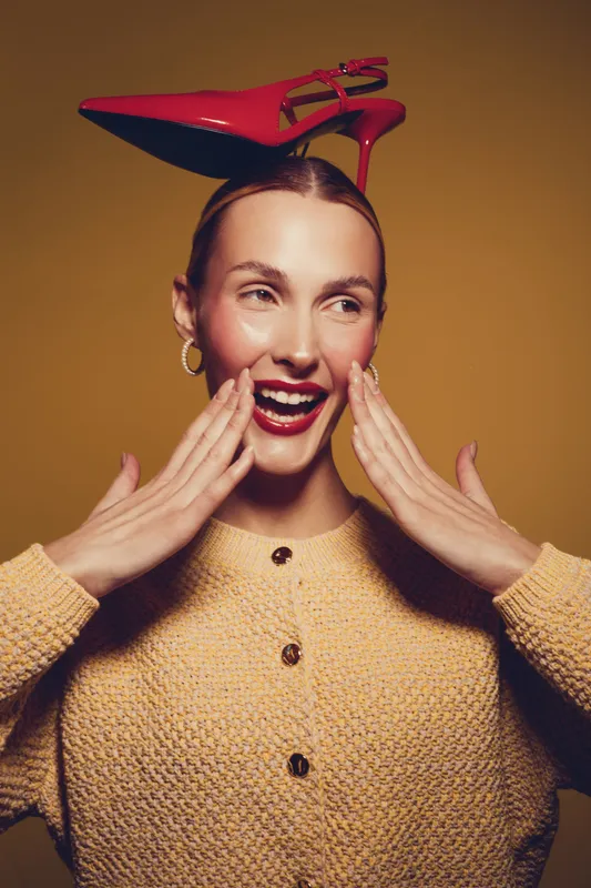 Smiling woman in beige sweater holds red high heel shoe on head, posing against yellow background. Concept for fashion, style, fun, and creative portrait with bright colors and joyful mood.