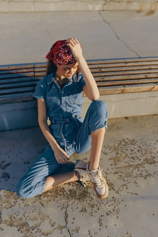young woman in denim jumpsuit with red bandana sitting on concrete pavement in sunlight outdoors with casual sneakers on a bright day