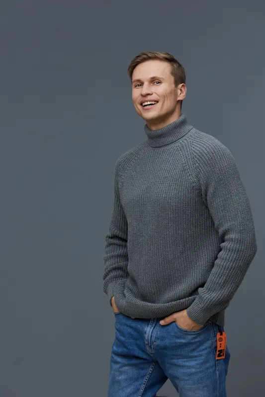 Smiling man wearing gray turtleneck sweater and blue jeans posing in front of plain gray background studio portrait cheerful expression casual style people concept