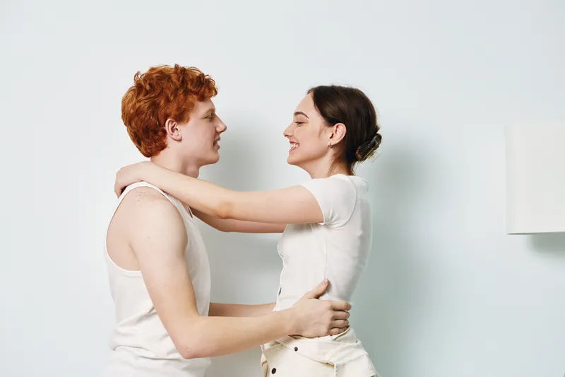 Couple embracing and smiling indoors, young man and woman in casual clothing with affectionate gaze and happiness in bright room with white walls and lamp in background.