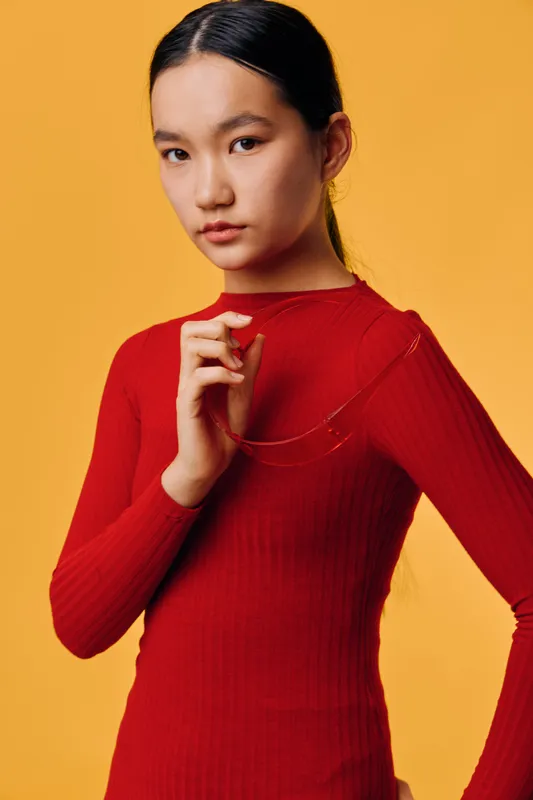 Young woman with black hair in red long sleeve top standing against orange background looking serious thoughtful and elegant with hand near chest and minimal makeup.