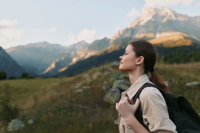 A woman hiking in the mountains with a backpack, outdoors, sunlight on her face, capturing a moment of exploration, solitude, and adventure amid vast scenery