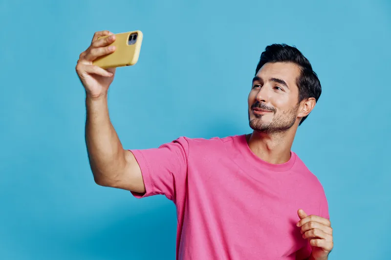 Happy smiling young man taking a selfie with mobile phone, wearing a bright pink t-shirt, isolated on vivid blue background, casual lifestyle concept.