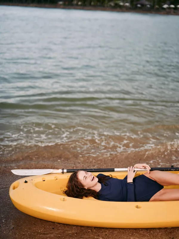 A woman lies inside a yellow kayak on a sandy shore, paddle resting across her chest, sunlit water gliding nearby, capturing a calm, warm daylight moment.