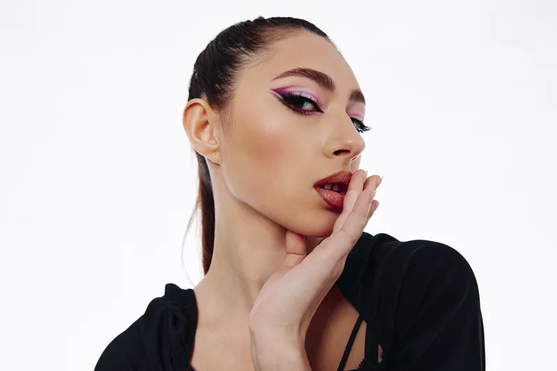 Creative beauty portrait of a young woman with pink glitter eye makeup and flowing long hair posing with hand near face on white background in studio. Fashion concept.