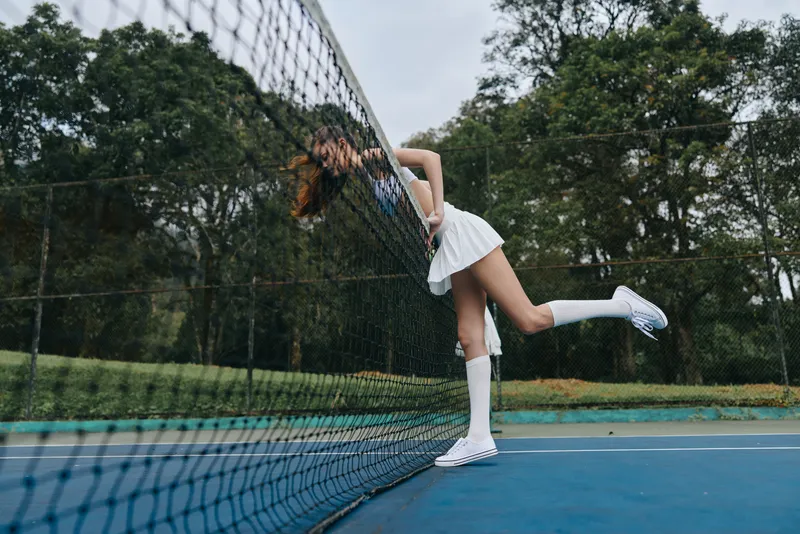 Young female tennis player in a white dress, posing playfully behind a net, conveying joy and energy in a vibrant outdoor setting