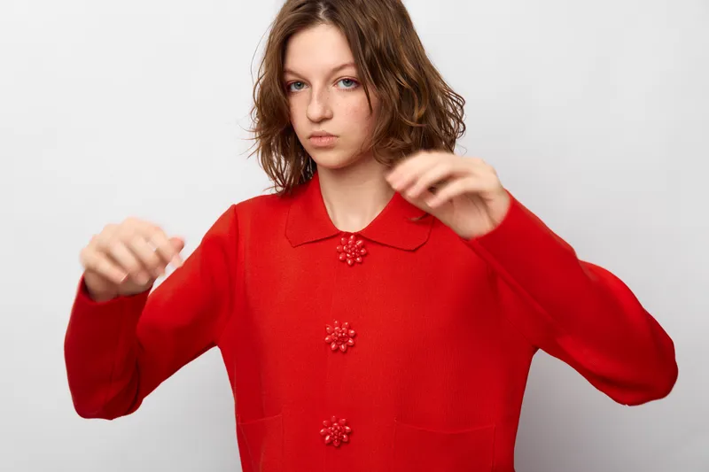 Young woman with curly hair wearing a red sweater poses with raised hands on a white background. Studio portrait with casual style and natural expression, concept of fashion and youth.