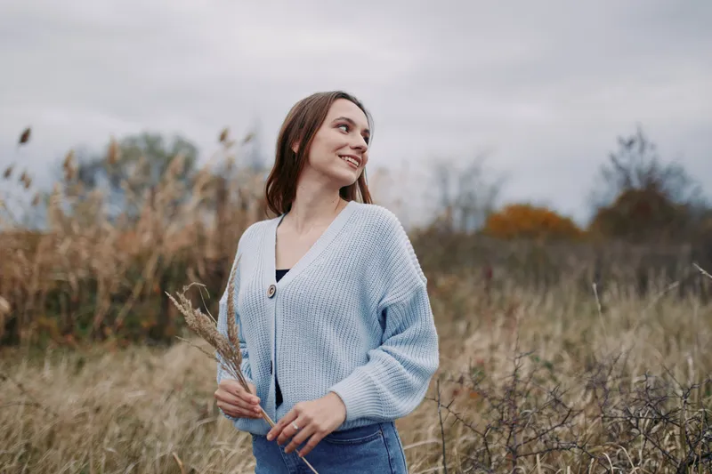 Candid portrait of a young woman standing in a wild meadow, holding dried grasses and smiling naturally in a soft knit cardigan under an overcast sky, her relaxed posture and honest expression convey
