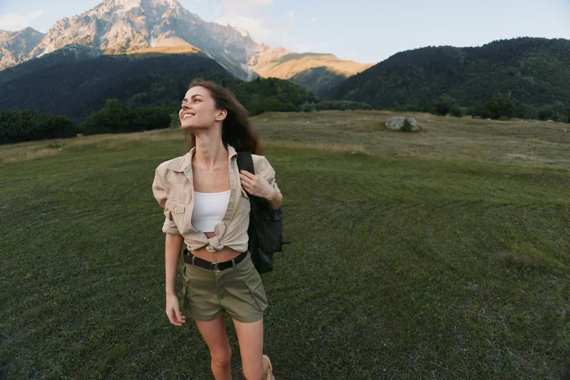 Young woman enjoys a sunny hike in a grassy field with distant mountains, backpack slung over one shoulder as she gazes upward with a smile.