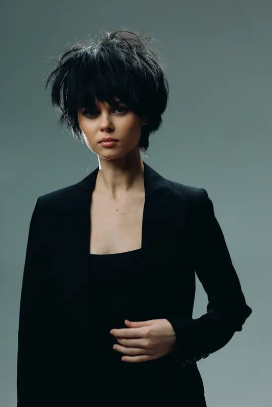 Young woman with short black hair wearing a dark blazer posing confidently with a neutral background and moody lighting.