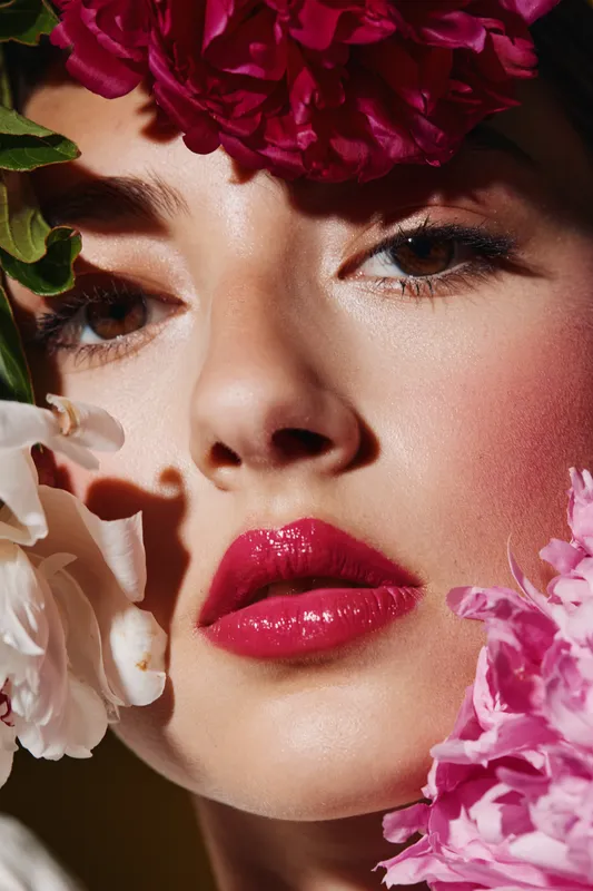 Close-up of a woman with glossy red lips and flawless skin framed by pink and white flowers. Fashion beauty portrait with creative vibrant hairstyling, makeup, and floral elements.