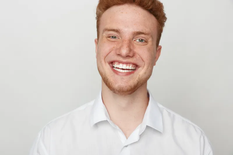 Smiling young man with red hair and freckles wears white shirt, showing bright teeth and happy expression on light grey background. Concept of joy, confidence, and positive emotions.
