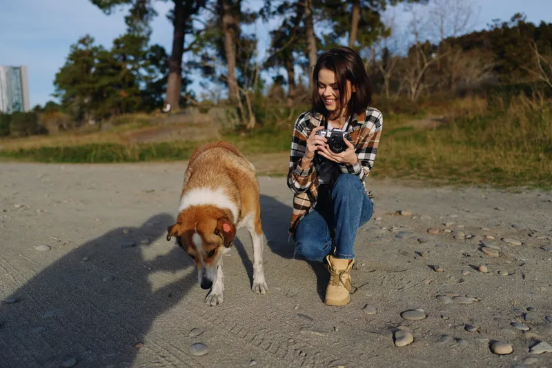 Woman with camera kneels on a sandy beach with her dog, capturing a candid outdoor moment in casual wear, plaid shirt and boots, under warm sunlight.