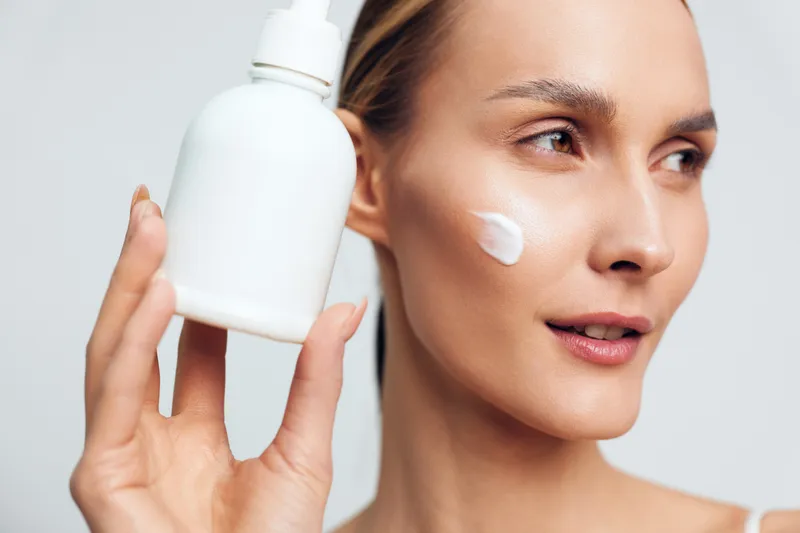A young woman with clear skin holds a white cosmetic bottle near her face with moisturizer cream applied on her cheek. Concept for skincare, beauty, and facial care routine.