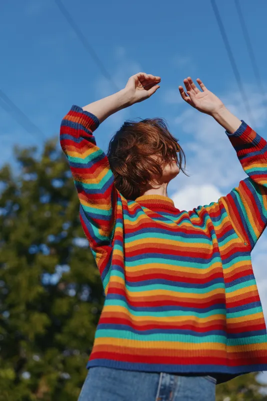 Person wearing rainbow sweater with arms raised outdoors under blue sky, portrait from low angle showing motion and expressive pose, casual jeans and textured hair in daylight.