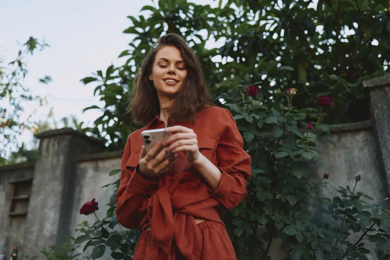 Woman outdoors wearing a red dress, smiling as she checks her phone, candid lifestyle moment amid lush garden greenery and soft natural light warm atmosphere and casual effortless vibe