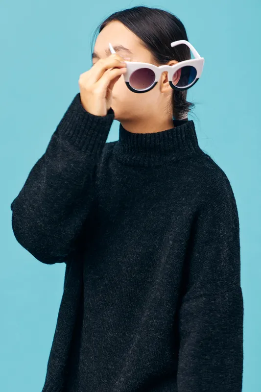 Woman wearing black sweater adjusting white oversized sunglasses on face with blue background. Casual fashion, hairstyle, accessory and stylish outfit for youth and modern look in studio.