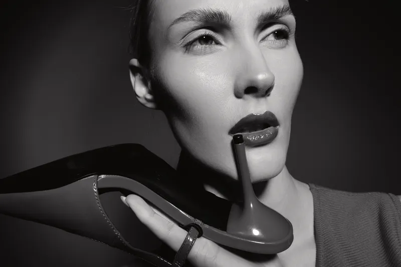 Closeup of a white woman with pale skin holding a high heel near her lips. Monochrome portrait with dramatic lighting and fashion concept.