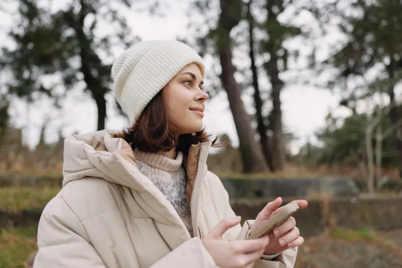 Woman outdoors in a warm coat and beanie using a smartphone, candid lifestyle portrait in a wintry park with soft natural light and calm urban vibe