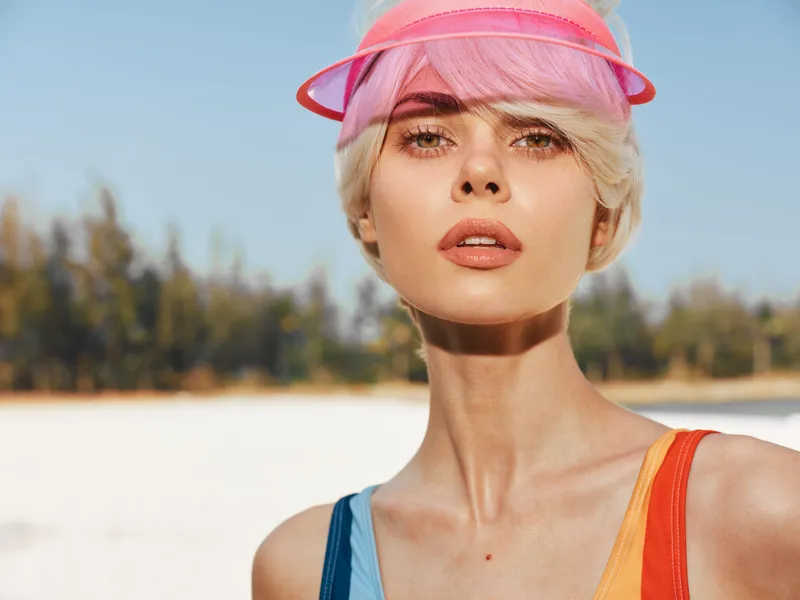 Sunlit portrait of a stylish woman at the beach wearing a vibrant swimsuit and pink visor, conveying bold summer confidence, glow, and a modern, carefree mood under clear light.