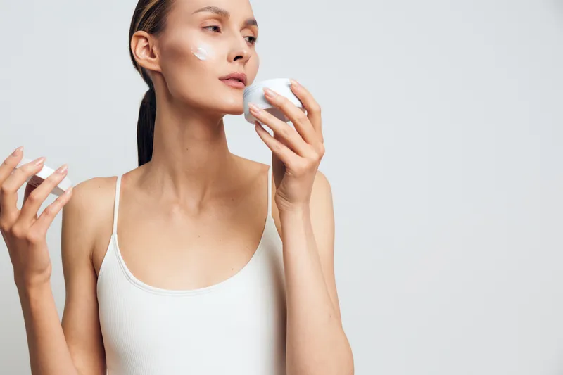 A young woman applies skincare cream to her face in a bright studio, capturing a quiet moment of self care and a calm daily routine.