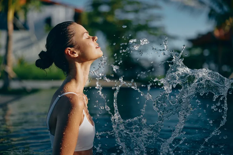 A young woman with wet hair enjoys fresh water splashing on her face while standing in a pool outdoors during sunny day. Concept for relaxation, summer lifestyle, and self care.