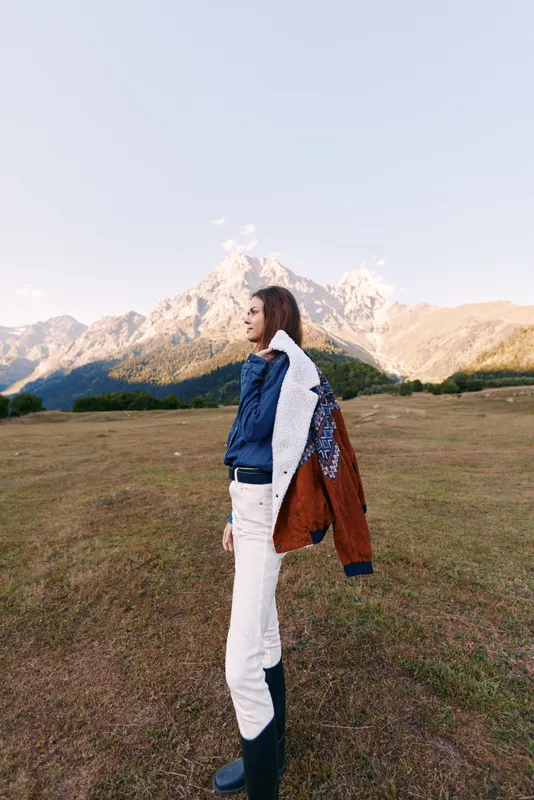 Woman in jacket standing in a mountain field, landscape portrait with peaks in the distance, outdoors scene showing casual fashion, boots and white pants in open meadow.