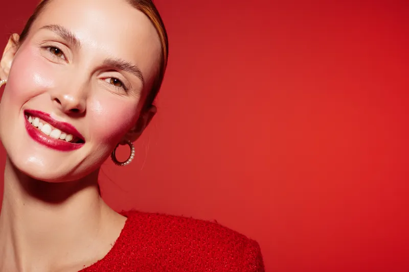 Smiling woman with red lipstick and earrings poses against red background wearing red textured top. Concept of beauty, fashion, makeup, and confident female portrait.