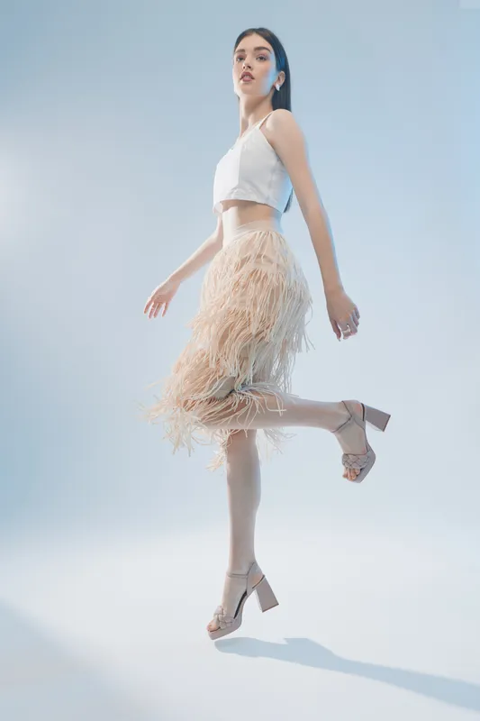 Fashionable woman in a white top and beige fringe skirt poses mid-air on a light background. Concept of beauty, fashion, elegance, and modern style for editorial or commercial use.