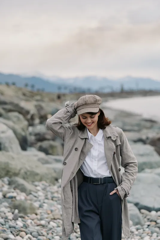 Young woman in gray coat and hat walks on rocky beach with mountains in background. She smiles and enjoys a cool, cloudy day near the sea. Casual autumn outfit and nature scene.