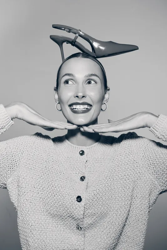 Young Caucasian woman with bright smile poses with high heel shoe on head, wearing knitted sweater. Creative concept for fashion, joy, and playful portrait in studio.