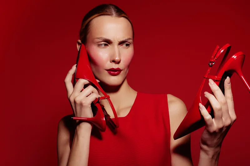 Woman in red dress holds two red high heels, looking confused against red background. Concept of fashion choice, style dilemma, and footwear selection in studio lighting.