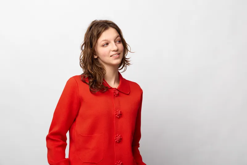 Young woman in red jacket looks sideways with slight smile on white background. Casual style, natural expression, and simple studio portrait concept for fashion or lifestyle.
