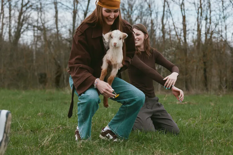 Two women share a joyful moment outdoors with a small goat kid, playing in a grassy meadow, natural light, casual outfits, warm mood, candid lifestyle scene, fresh air, rustic charm, everyday