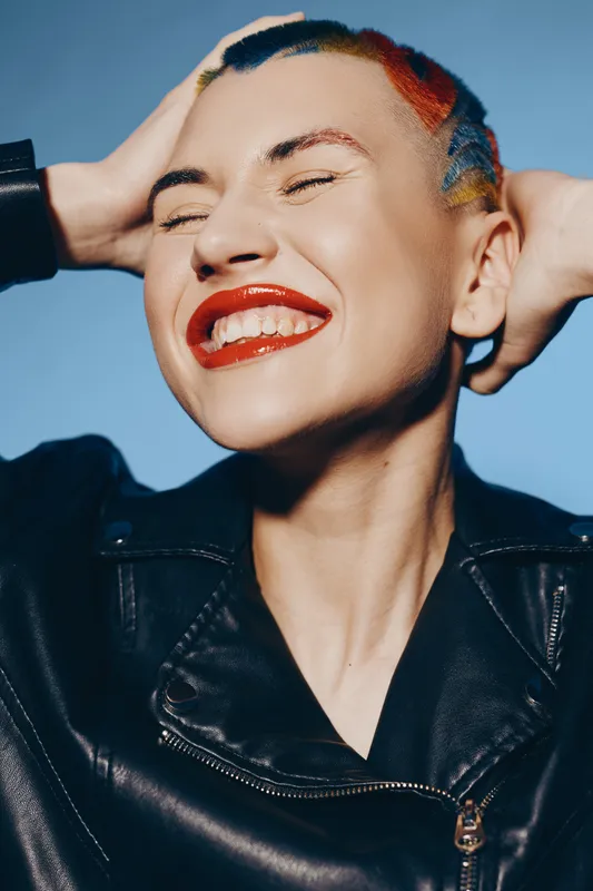 Smiling woman with colorful short hair and red lipstick wearing black leather jacket, expressing joy and confidence against a plain blue background.