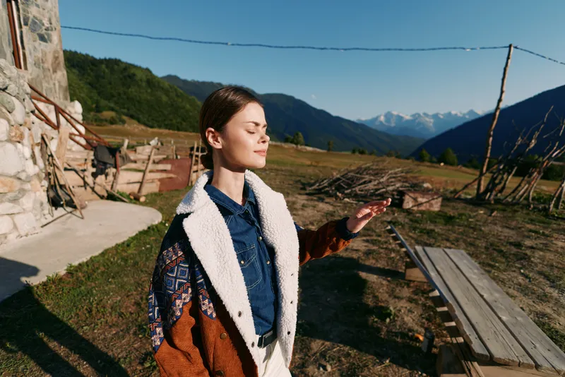 Woman standing outdoors in countryside, portrait with eyes closed enjoying sunlight and nature near rustic cabin and wooden fence, cozy coat and serene mountain landscape background