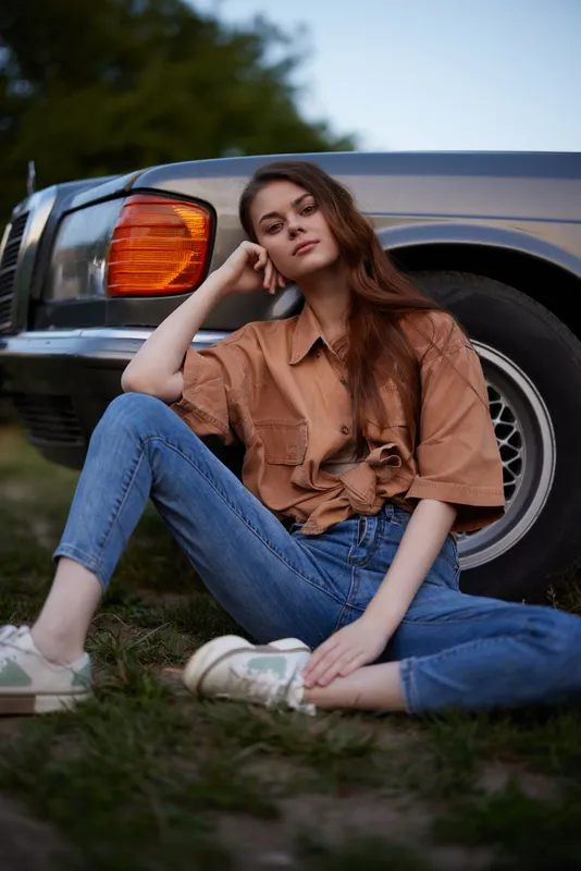 Young woman relaxing outdoors near a vintage car, wearing a brown shirt and blue jeans, embodying a carefree summer vibe with natural beauty.