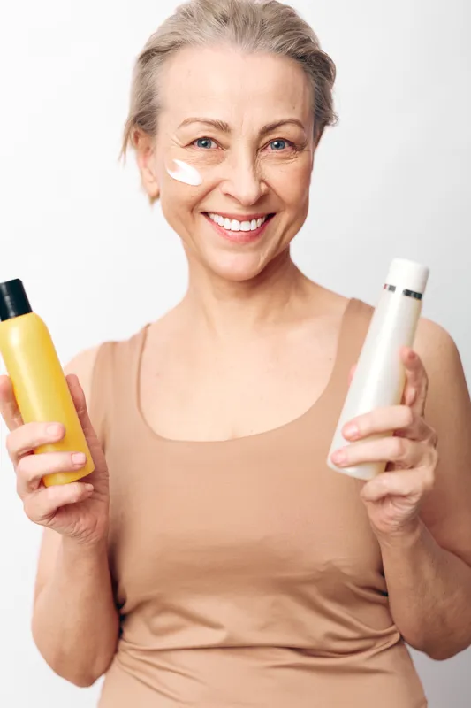 Smiling mature woman in beige tank top holds two cosmetic bottles with cream on her cheek. Concept of skincare routine, self-care, beauty, and healthy aging in bright studio setting.
