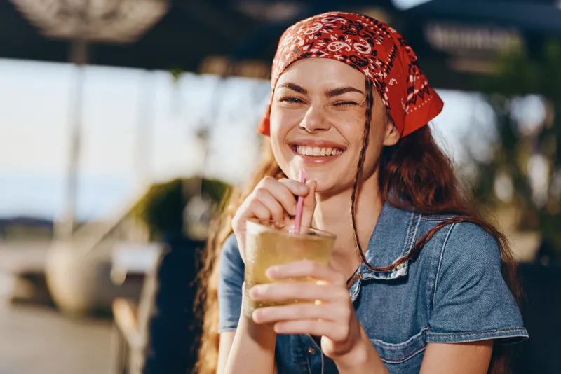 smiling woman in modern boho-western denim street style drinking beverage outdoors, film color, casual joyful mood under sunlight with natural blurred background