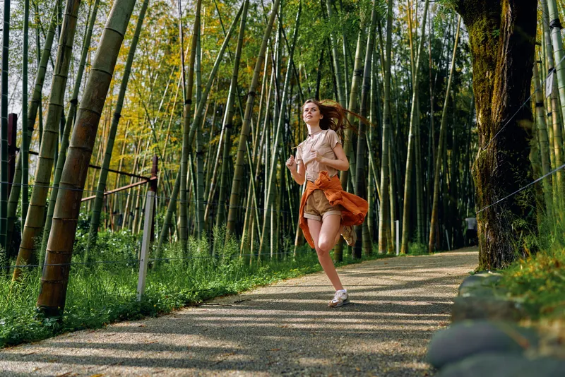 Woman running in a bamboo forest path, dress flowing and joyful against green stalks, sunlight and shade on a gravel trail, active outdoor scene conveying freedom and movement.