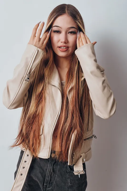 Young woman with long hair wearing beige jacket and black jeans posing indoors against white background. Fashion style, casual outfit and portrait of confident female model with natural makeup.