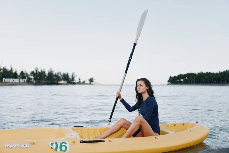Young woman paddling a yellow kayak on calm water during a clear day, surrounded by distant trees and a serene sky.
