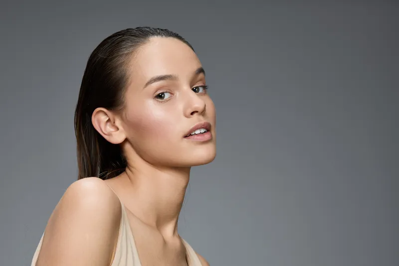 Elegant young woman with natural makeup and long straight hair posing against a gray background, showcasing beauty and confidence in a minimalist style