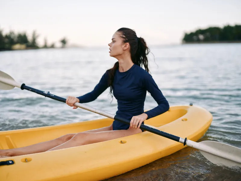 Young woman kayaking on calm water wearing navy shirt, paddling yellow kayak during outdoor activity, peaceful nature background.
