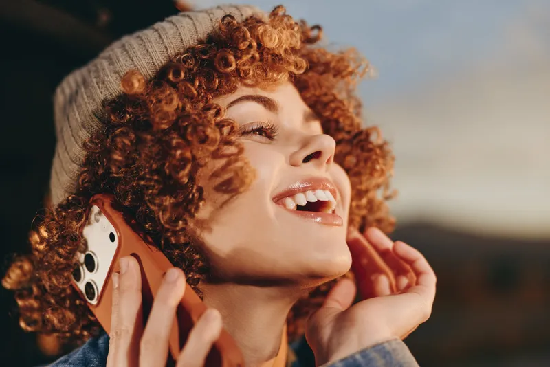 Woman lifestyle closeup curly hair smiling holding smartphone to ear outdoors, soft light shallow depth of field, casual jacket, friendly communication and everyday tech use today